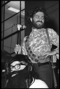 Jerry Rubin, with bandolier and toy assault rifle slung over his shoulder, addressing the audience at the microphone