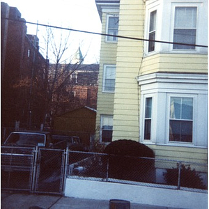 Partial view of the exterior of a triple-decker yellow house with white trim, with a pickup truck and car parked in the adjacent driveway (on left), Roxbury, Mass