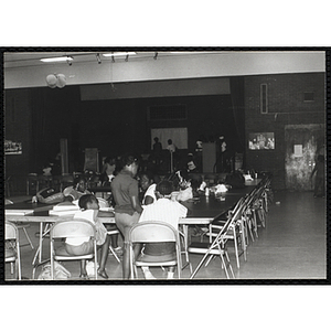 Several Roxbury Boys and Girls Club members sit and stand around tables in an auditorium while others stand on the stage in the distance