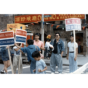 Three people stand in the street holding Mel King campaign signs during the August Moon Festival in Boston's Chinatown