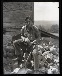 Lowell Ames Norris, seated on rocks with a shovel, holding a human skull