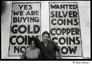 Lissa Matross and Harvey Wasserman standing in front of a sign at a curio store: 'Yes we are buying gold coins...'