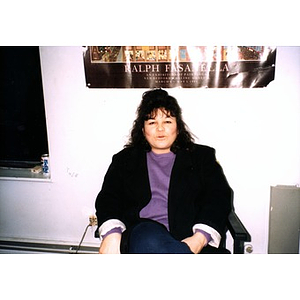 Woman in a purple shirt sitting beneath a poster that advertises an exhibition of works by Ralph Fasanella.