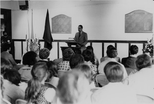 Randolph W. Bromery standing indoors, behind podium, before an audience