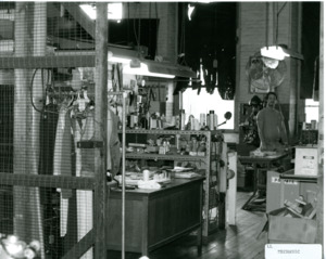 Photograph of a man standing in a room with tools and supplies, [1982-1983].