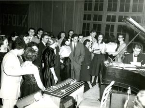 Students in formal wear gathered around man playing piano at a Suffolk University dance, 1952