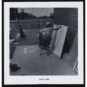 Three boys paint on plywood propped against a brick wall as another boy looks on during Tom Sawyer Day