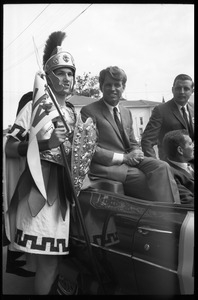 Robert F. Kennedy seated in an open car next to a high school student in Trojan costume at the Turkey Day parade