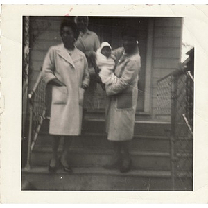 Three women, one holding an infant, stand on the front steps of a house