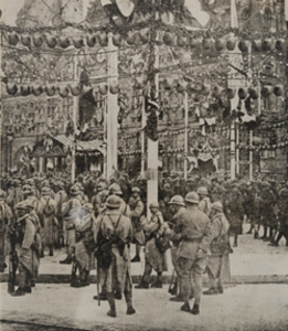 Street-level view of a large crowd of French soldiers standing on a city sidewalk, the street hung with flags and bunting, Paris