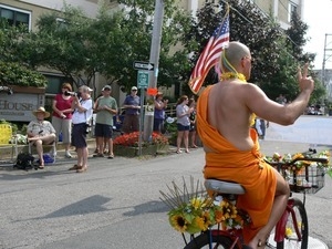 Man with shaved head and dressed in a saffron robe bicycling in the parade: Provincetown Carnival parade