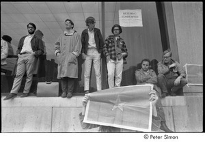 MIT war research demonstration: demonstrators standing by a window outside a building, woman (possibly Lacey Mason) in foreground, holding NLF flag