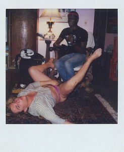 A Photograph of Marsha P. Johnson Sitting in a Chair While Paulina Lays on her Back on the Floor with Her Legs Up