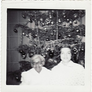 An elderly housekeeper sits beside a Christmas tree