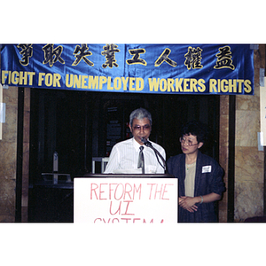 An unidentified man wearing a shirt and tie speaks at an event at the Massachusetts State House for unemployment insurance for workers, with Suzanne Lee standing on right