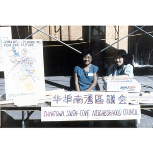 Two women sit behind an information table outside on a sidewalk for the Chinatown South Cove Neighborhood Council