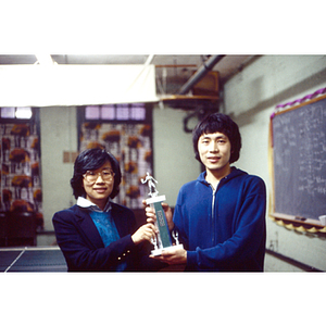 Man stands next to Suzanne Lee, after winning the table tennis competition, both of them holding the trophy