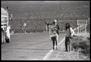 Activists flashing a peace sign at a passing truck by the road near entrance to the Nevada Test Site, holding a sign reading 'Stop testing now': Nevada Test Site peace encampment