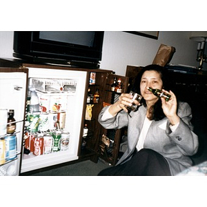 Clara Garcia sitting on the floor in front of a hotel mini bar pretending to pour a small bottle of liquor into a glass.