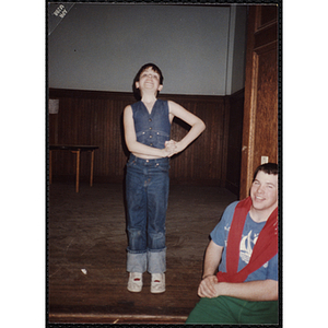 A young boy stands on a stage with his hands clasped above his left waist while a young man sits in front of him and smiles for the camera at the Charlestown Boys and Girls Club