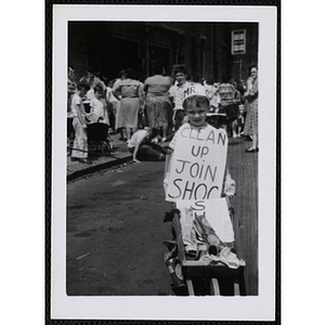 A boy carries "Clean up join SHOC" placard with small wheelbarrow at a carnival