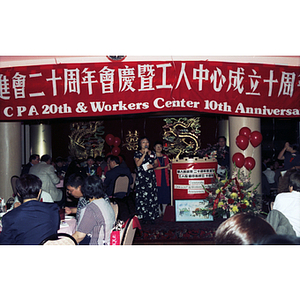 Lydia Lowe stands behind a woman addressing the crowd at a restaurant for Chinese Progressive Association's 20th Anniversary Celebration