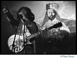 Bob Marley in concert, exhorting the audience while standing in front of a large banner with a portrait of Haile Selaisse