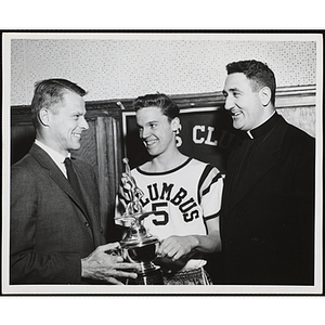 Richard Harte, Jr. presents a trophy to a young man in a basketball uniform while a priest looks on