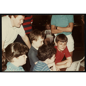 A Group of children gathering around a computer keyboard while an unidentified adult looks on from behind