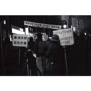 Rear view of a crowd listening to a woman speak on a stage at an evening demonstration held in Chinatown against the encroaching Combat Zone, the infamous red-light district in Boston