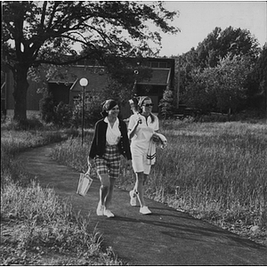 Two female students with tennis rackets walking up a paved path