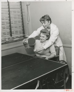 Mrs. Frances Marsala seated in her wheelchair playing ping pong assisted by attendant