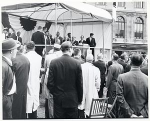 Mayor John F. Collins seated on tented platform outdoors and surrounded by unidentified men