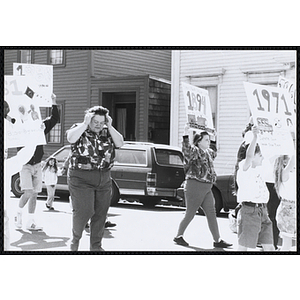 A woman puts her hands on her head while walking along the street with the children holding up their signs during the Boys and Girls Clubs of Boston 100th Anniversary Celebration Parade