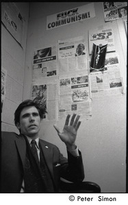 Richard Hughes: close-up portrait of draft resister gesturing, seated beneath wall plastered with BU News