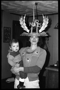 Nina Keller, making a silly face, wearing a winged No Nukes headdress, holding a baby, Montague Farm commune