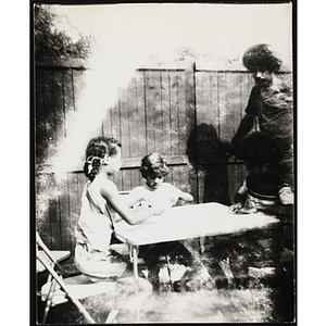 Three children play cards at a table outdoors as a man looks on