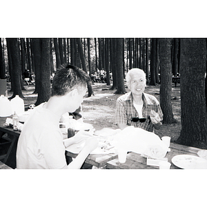 Young man sits across a picnic table from another man who is eating a piece of watermelon