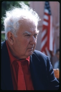 Alexander Calder: three-quarter profile bust portrait in a red tie, with an American flag in the background