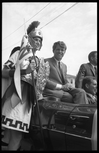 Robert F. Kennedy seated in an open car next to a high school student in Trojan costume at the Turkey Day parade