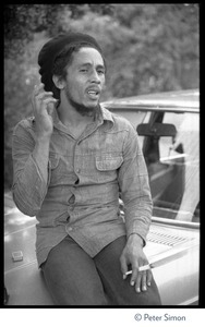 Bob Marley: three-quarter length portrait, seated on the hood of a car, smoking a blunt