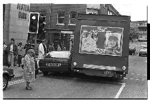 Old woman crosses street beside Coca Cola sign printed on the back of a caravan in Downpatrick