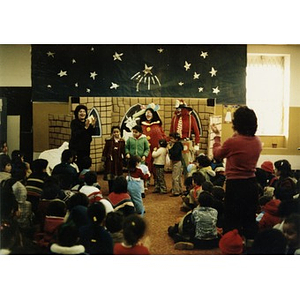 Audience watches four little girls and two clowns onstage at a Three Kings' Day celebration at La Alianza Hispana, Roxbury, Mass., while two women stand to their feet and applaud