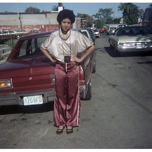 Full-length portrait of a Hispanic American woman, facing front, wearing athletic pants, standing in front of a red car that is parked in the street at a Latino street festival.
