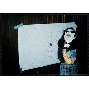 A child wearing a mask stands next to a poster of a spider in a web at a Halloween event