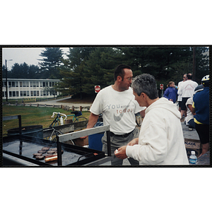 A man and a woman attend to a grill in a parking lot during The Partnership Ride