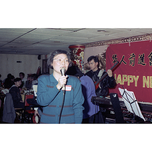 Woman speaks from a microphone while a band sets up during a Chinese Progressive Association celebration of the Chinese New Year