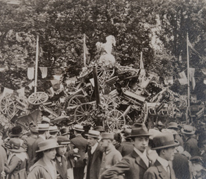 Close-up view of civilians in front of a large pile of captured German small artillery covered with wreathes, topped with a statue of an oversized rooster and flags from different countries hanging behind, 1919
