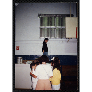 A group of boys and girls, facing away from the camera, stand together while a girl walks past in the distance in a Boys & Girls Club gymnasium