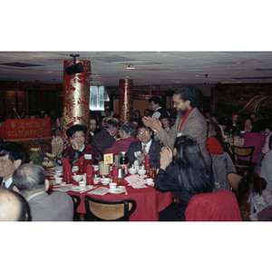 Rep. Byron Rushing stands facing Suzanne Lee at a restaurant table while he and others applaud